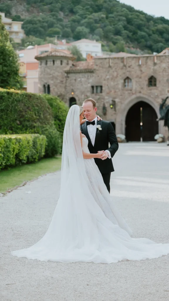 Two people in wedding attire dance near a historic stone building, surrounded by greenery and distant hills. The setting is elegant and picturesque.