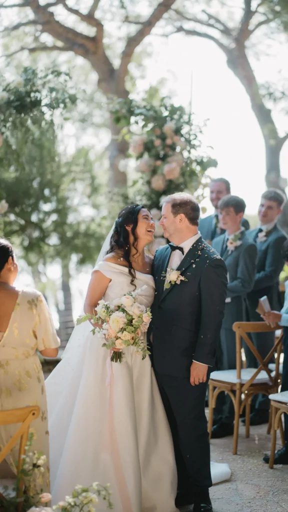 A couple stands under floral arrangements during an outdoor wedding ceremony, surrounded by people and wooded scenery.