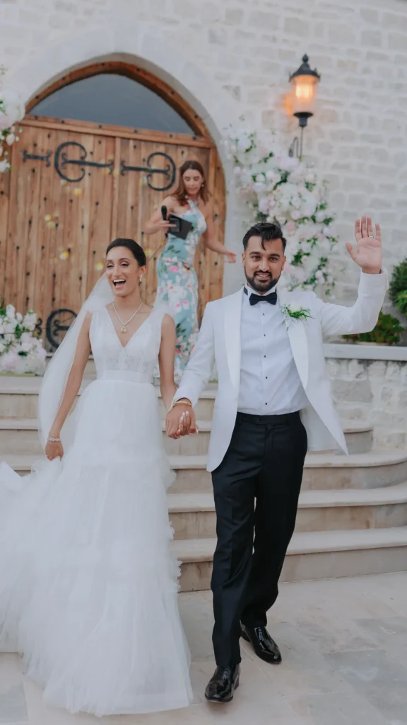 A couple in wedding attire joyfully exits a stone building with wooden doors, accompanied by a person holding a microphone.