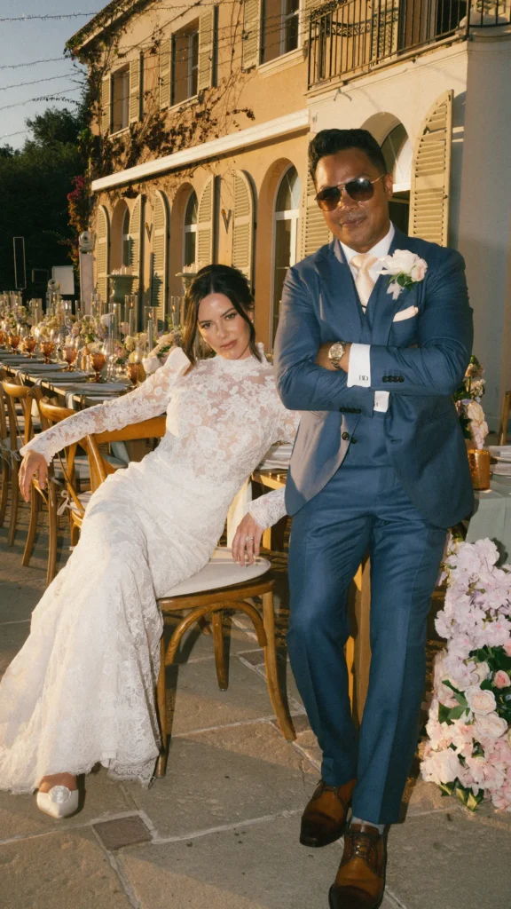 A person in a lace dress sits on a chair outdoors, beside another in a suit, near a decorated table and villa.